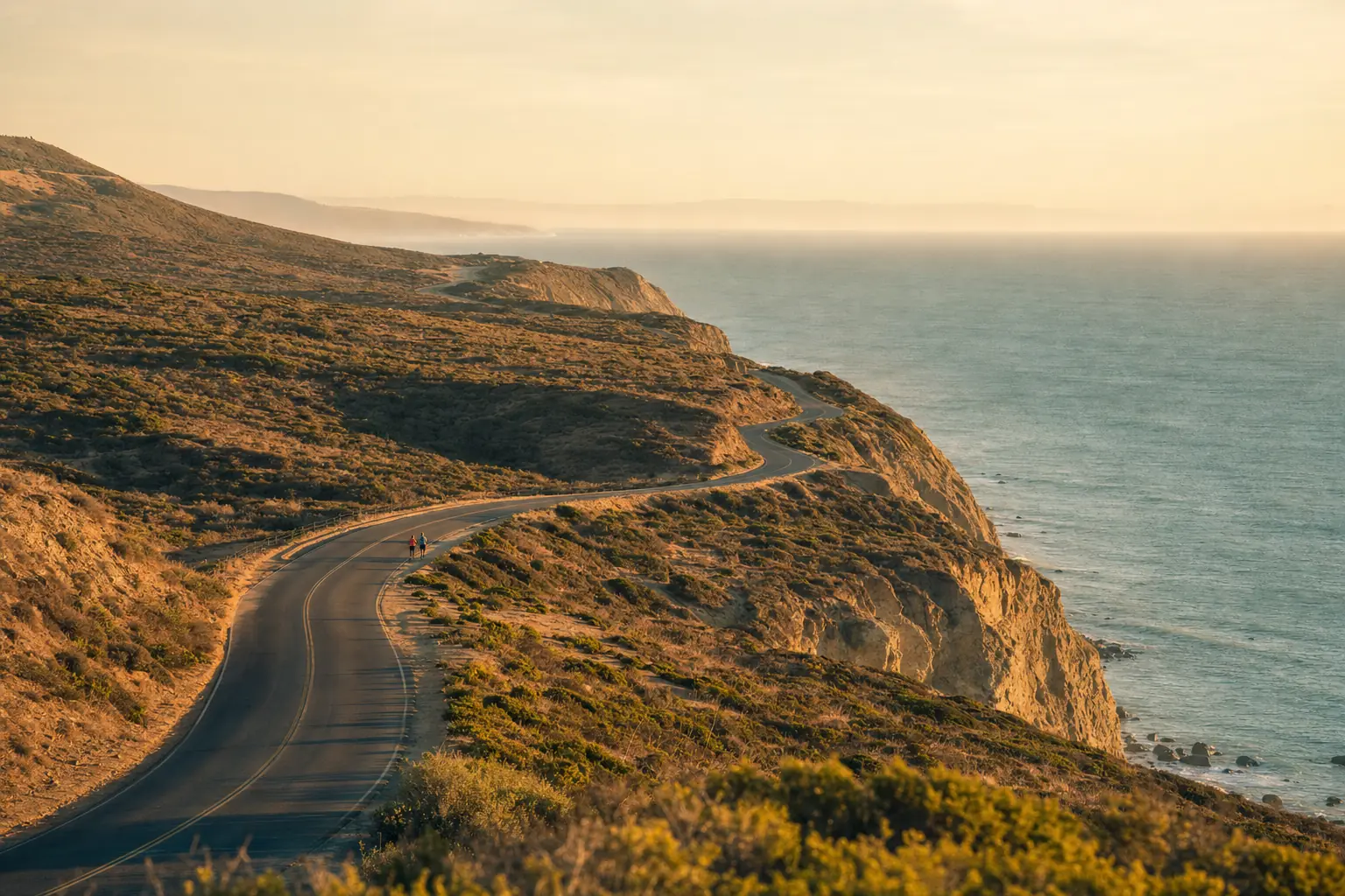 Two distant runners on a coastal road winding along golden cliffs above the sea at sunset